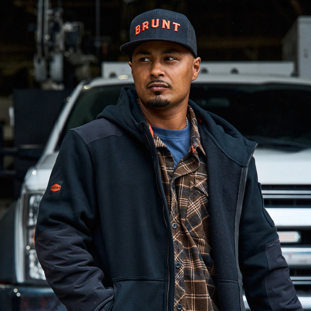 Man wearing flat brim work hat with built in pencil holder and BRUNT logo in black and orange on jobsite