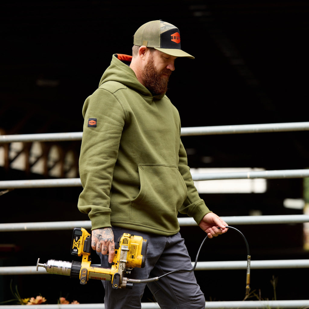 Man wearing flat brim work hat with built in pencil holder and BRUNT logo in army green camo carrying power tool
